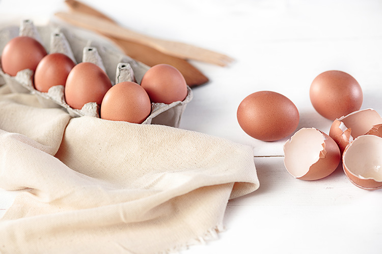 fünf Eier in einem offenen Eierkarton, rechts daneben auf der weißen Tischpaltte liegen zwei ganze und zwei aufgeschlagenen Eier