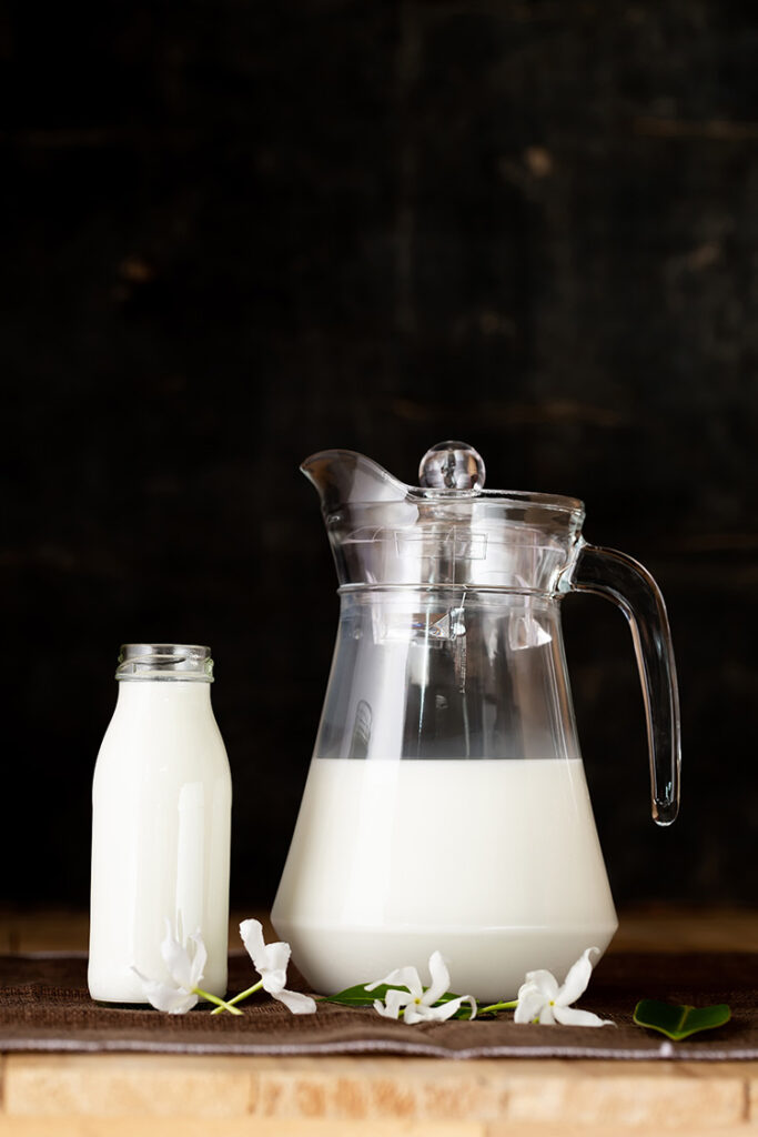 eine Glaskaraffe mit Milch und eine gefüllte Milchflasche aus Glas stehen auf einer Tischplatte aus Holz